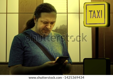 A young man in casual clothes looks surprised and displeased at a mobile phone screen at dusk near the "TAXI" sign