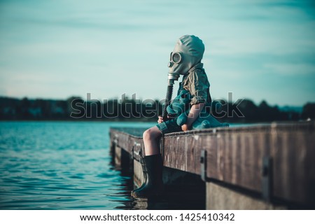 The child in the gas mask is sitting on the platform at the lake. Environment pollution. Coronovirus epidemic. Royalty-Free Stock Photo #1425410342