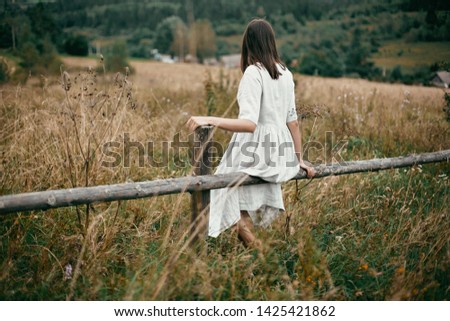 Stylish girl in linen dress sitting on aged wooden fence among herbs and wildflowers, looking at field. Boho woman relaxing in countryside, simple slow life style.  Atmospheric image Royalty-Free Stock Photo #1425421862