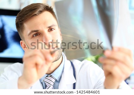Portrait of serious physician examining attentively sciagram. Concentrated doc holding sciagraph and pointing with pen to injured place. Medical treatment and health care concept. Blurred background