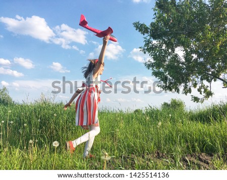 Girl red striped style dress. Landscape green background. Grass and tree. Child playing mini airplane. Natural texture background blue sky and white clouds. Mock up banner for text box. Sunlight.