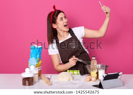 Chef cook confectioner or baker in brown apron, white t-shirt, red hairband, making cake at table, doing selfie on mobile phone, posing with open mouth isolated over pink background in studio.