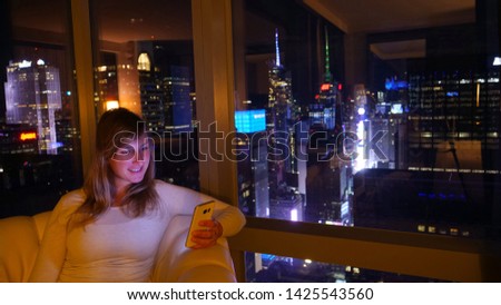 CLOSE UP: Cheerful Caucasian woman sitting on the couch and texting from her hotel room overlooking downtown NYC. Tourist girl browsing the internet on her smartphone while in room above Times Square