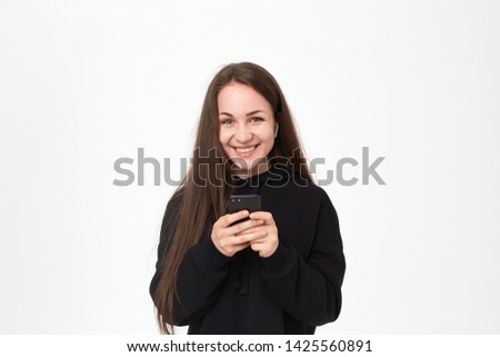 Studio shot of a beautiful young brunette woman with wireless headset and phone looking happy at the camera while standing over white background. Girl wears black hoodie.