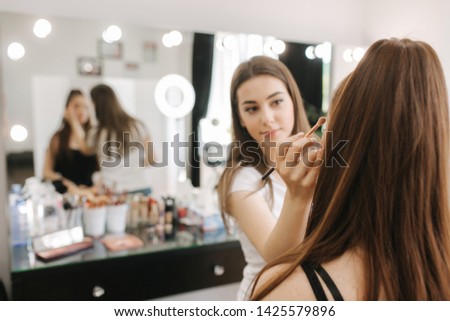 Beautiful femalemakeup artist doing makeup for a young redhead girl in a beauty salon sitting in front of a large mirror. Concept of preparation for the holiday and meeting