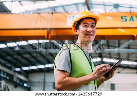 Young Asian male heavy industral worker holding a digital tablet inside manufacturing and factory site with crane behind