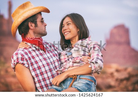 Lovers cowboy and cowgirl in love cute couple in USA backcountry landscape. Boyfriend wearing cowboy hat carrying Asian girlfriend seducing and flirting.
