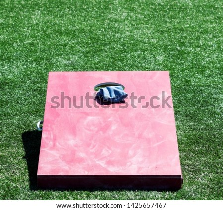 A hand painted homemade red cornhole game is on a green turf field with a blue bean bad in the hole.
