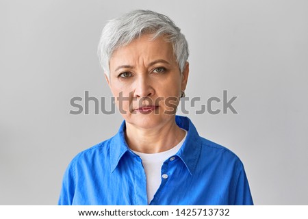 Horizontal picture of serious grumpy gray haired mature businesswoman in stylish blue shirt expressing negative emotions, frowning eyebrows, displeased with late employee or missed deadline