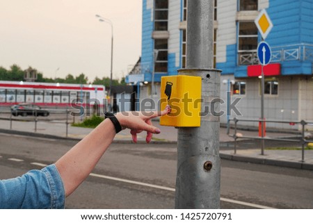 The hand of a girl with a clock presses the button of the pedestrian crossing. A hand sign indicates. The button is lit in red, modern electronic pedestrian crossing.                               