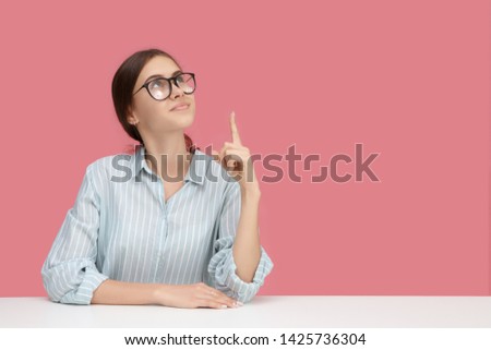 Creative mind, ideas, education and occupation concept. Picture of nerdy smart young Caucasian woman wearing blue shirt and eyeglasses posing at blank pink wall, pointing index finger upwards