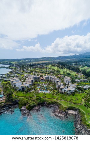 An aerial panoramic view of Kapalua Bay, Namalu Bay, and the surrounding resorts in Maui, Hawaii. Showcases the clear, blue, ocean sea, white, sandy beach, bright sky, and surrounding coral reef.