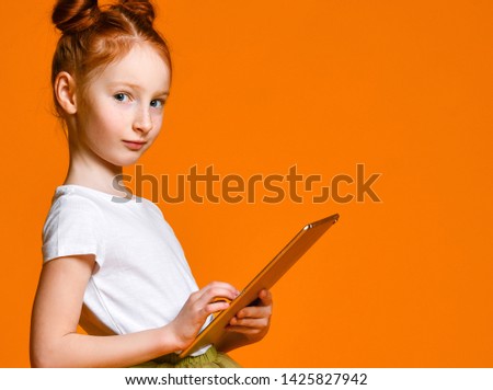 Cute red-haired girl school girl holding a tablet and looks into it. Side view. Child playing game, surfing or watching cartoon