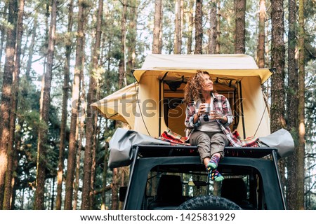 Happy hipster traveler people enjoying the alternative vacation - car with tent on the roof and beautiful young woman sit down outside looking at the forest around - outdoor activity Royalty-Free Stock Photo #1425831929