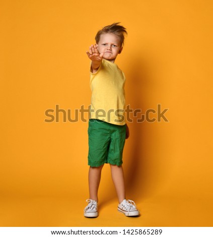 Serious-looking young man kid boy in yellow t-shirt and green shorts shows stop sign gesture on yellow background