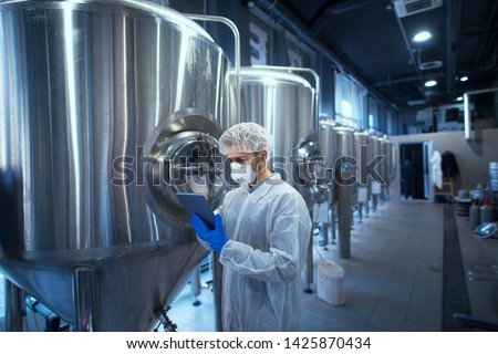 Factory worker technologist in protective uniform with hairnet and mask controlling food production on tablet computer.