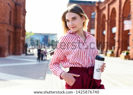 Portrait of beautiful girl dressed in red shirt, with take away coffee in cup. Blurred background of city center, building with red brick walls.