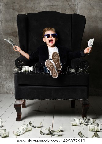 Happy and shoutting arrogant rich kid boy millionaire sits with a bundle of money dollars cash in big luxury armchair while bills falling down from the sky. Money rain