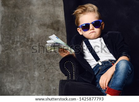 Happy and shoutting arrogant rich kid boy millionaire sits with a bundle of money dollars cash in big luxury armchair while bills falling down from the sky. Money rain Royalty-Free Stock Photo #1425877793