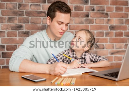 Serious concentrated young dad in sweater sitting at table and drawing picture with daughter, little girl looking at father