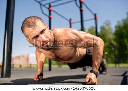 Young muscular bodybuilder standing on fists over ground while doing push-ups during workout outdoors