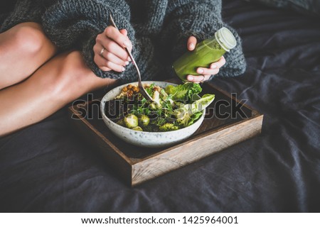 Healthy dinner, lunch setting. Vegan superbowl or Buddha bowl with hummus, vegetable, salad, beans, couscous and avocado, smoothie on tray and woman in warm sweater eating in bed Royalty-Free Stock Photo #1425964001