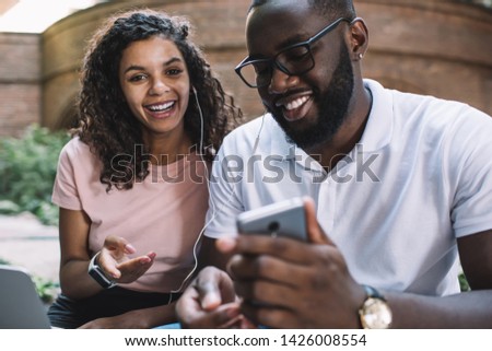 Portrait of excited black hipster girl with curly hair smiling at camera while spending spare time with positive boyfriend listening music togetherness from smartphone application via headphones