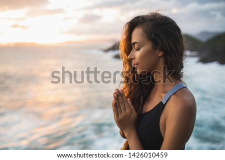Young woman praying and meditating alone at sunset with beautiful ocean and mountain view. Self-analysis and soul-searching. Spiritual and emotional concept. Introspection and soul healing. Royalty-Free Stock Photo #1426010459