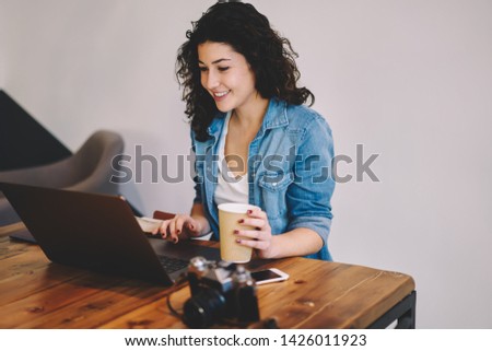 Happy hipster girl dressed in casual clothing watching interesting video on website via laptop computer connected to wifi, cheerful female freelancer enjoying caffeine beverage while working online
