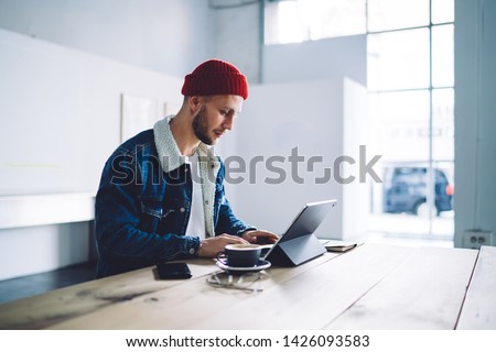 Trendy dressed male blogger editing media pictures via graphic application on modern touch pad connected public wifi internet for working remotely, Caucasian hipster guy checking analytic of blog