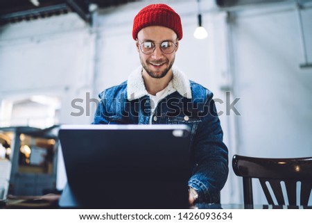 Young positive Caucasian student checking mail message by electronic gadget connected to wifi, happy hipster guy enjoying free time with touch pad for chatting sitting indoors and waiting friend