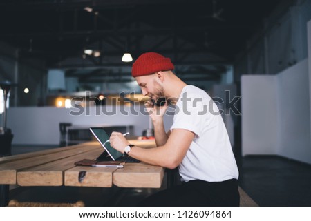 Side view of millennial graphic designer tapping modern tablet with stylus while working at wooden desk in coworking space, successful man making cellphone conversation for discussing project details