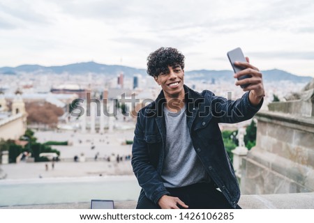 Portrait of cheerful Latino male traveller resting during Spanish vacations to Barcelona clicking selfie pictures on Montjuic area, millennial man photographing himself with cityscape on background