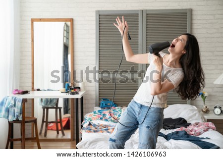 funny girl singing with hair dryer as microphone after dries long hair. beautiful asian lady relax having fun playing in bedroom with messy bed full of clothes in house on summer holiday hot outdoors