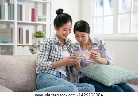 Two joyful young excited asian women friends having fun point on mobile phone screen while sitting on comfortable sofa at home. Female friendship chatter funny pictures on cellphone pleasant memory