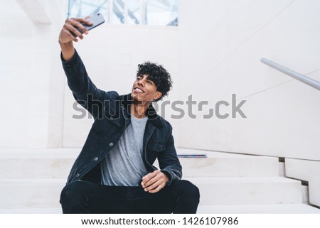 Cheerful Latino man with cellular gadget clicking selfie pictures on smartphone camera resting at city area, happy young male shooting video for blog in social networks using modern mobile phone