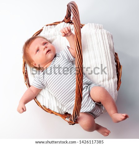Portrait of a newborn baby boy, age 1 month, in a striped bodysuit, red hair, blue eyes on white bedclothes in a wicker basket on a white isolated background. Family, love, children concept
