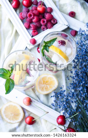 
Lemonade with lemon, raspberry, cherry and mint. On a light background in a stylish rustic with a bouquet of lavender, photo frames and scattered berries