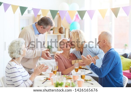 Curly-haired lady kissing birthday woman and saying wishes in ear while giving gift at birthday party