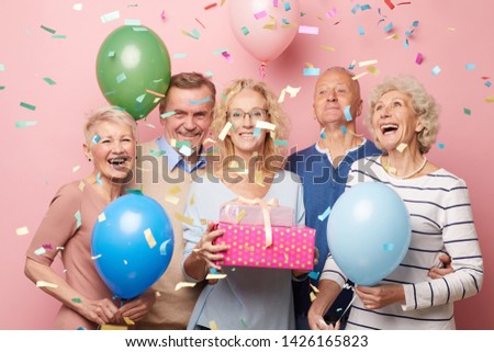 Cheerful excited mature friends in casual outfits standing against pink wall under falling confetti and having fun together at birthday party