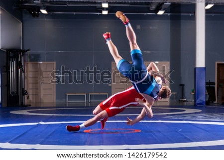 Two young men in blue and red wrestling tights are wrestlng and making a suplex wrestling on a wrestling carpet in the gym