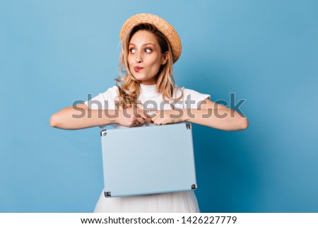 Curly woman looking slyly to side and posing with blue hand suitcase. Snapshot of a lady in white dress and straw hat