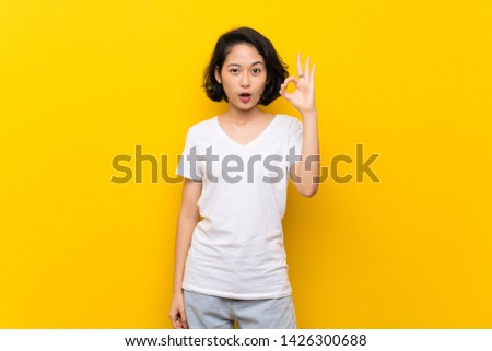 Asian young woman over isolated yellow wall surprised and showing ok sign