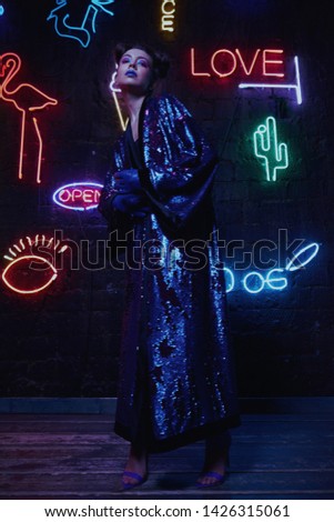 Cyberpunk style portrait of girl in futuristic purple bathrobe with glitter. She poses against wall of neon figures. Set is lit with magenta light. Clothes is oversized. Picture has dark noir tones.