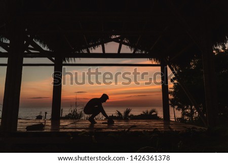View of man rolling out yoga mat and looking at beautiful orange, yellow and blue sunset over Montanita beach town from bamboo hut. Shot in Ecuador.