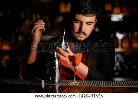 Professional male bartender spraying from the diffuser on the cocktail in the glass on the bar counter in the dark blurred background