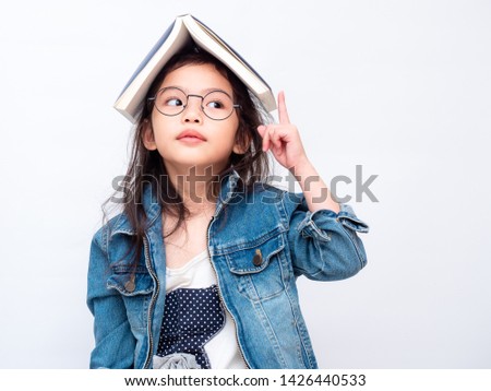 Asian little cute girl wearing glasses and put the book on head. Preschool lovely kid with the book covering on her head and pointing finger up. Learning and education of kid. Royalty-Free Stock Photo #1426440533