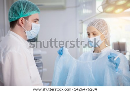 Surgical nurse helps the doctor to wear a bathrobe in the operating room, preparing for surgery.