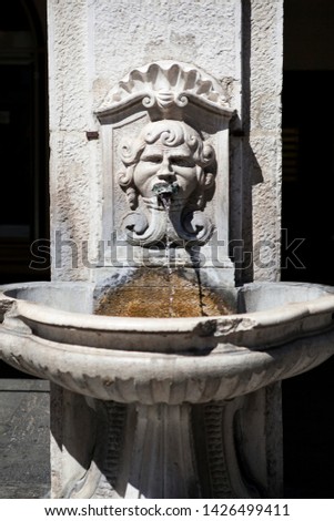 The antique old stone fountain with drinking water in the north of Italy, Lombardy, Brescia. Head of a man with long hair and a metal frog in his mouth a tap for water. Western Europe. Heritage.