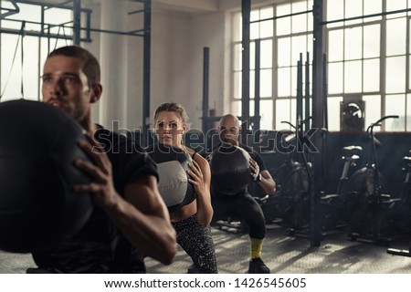 Group of three people doing squats while holding heavy medecine balls. Young men and fitness woman doing bodyweight exercises using weight ball. People in a row squatting with weighted balls at gym. Royalty-Free Stock Photo #1426545605